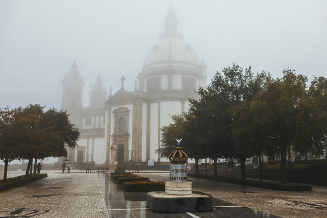 A grand Portuguese basilica with baroque dome looms through morning fog near Braga, a golden crown monument marking the centenary in the foreground.