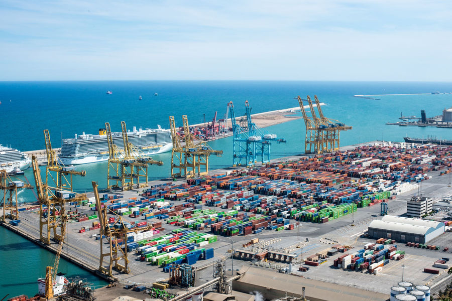 A vibrant aerial shot of Barcelona port featuring cranes, containers, and cruise ships on a sunny day.