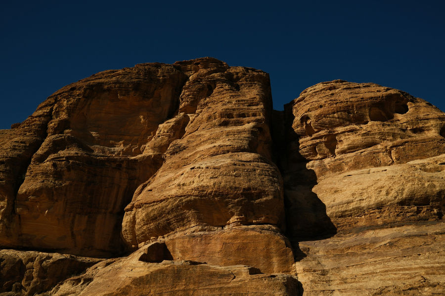 Dramatic lighting on a massive sandstone cliff face in Petra, highlighting the intricate horizontal sediment layers and deep shadows of the canyon.