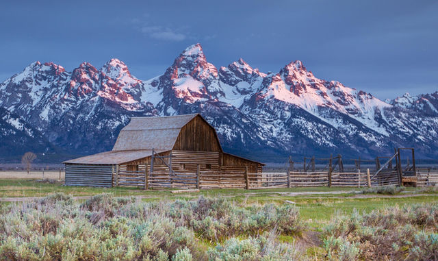 A wooden barn with snowy peaks in the background and green vegetation in the foreground