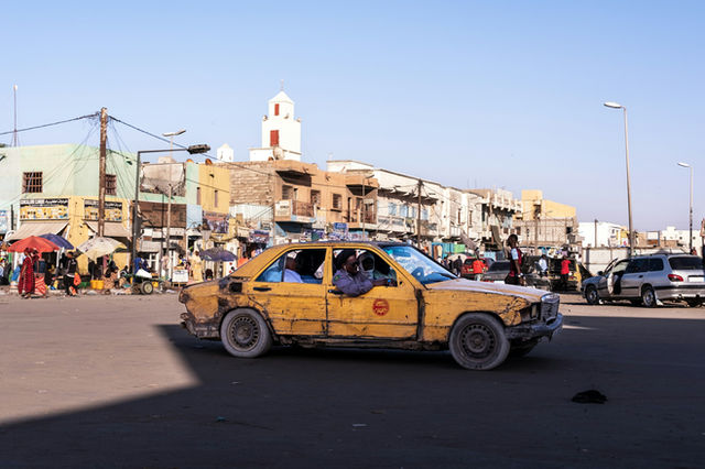 A worn yellow taxi drives through a bustling town square
