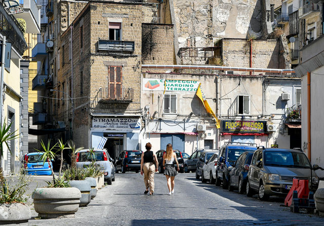 Two women walk down a narrow street lined with cars