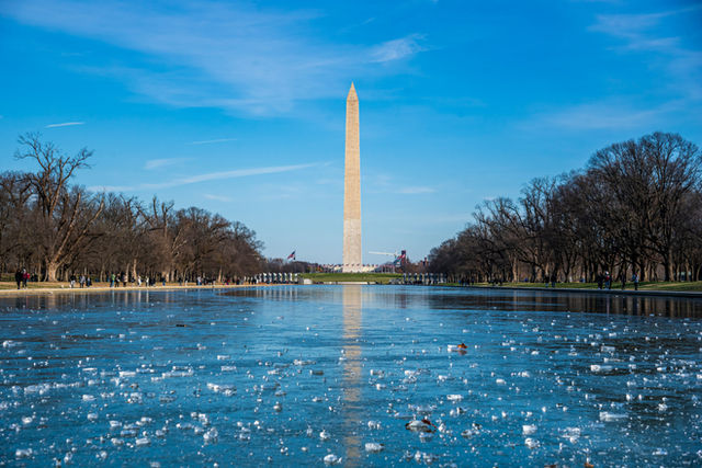 Photographed a winter view of the Washington Monument centered at the end of a partially frozen Reflecting Pool, framed by bare trees and pedestrians walking along the National Mall.