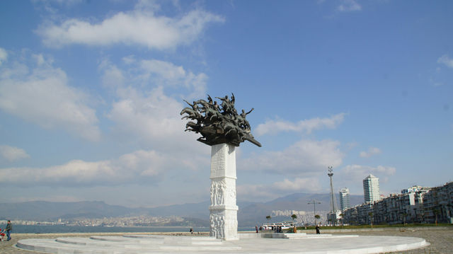 A tall white pedestal supports a dynamic sculpture of riders on horses. The monument stands near the waterfront with mountains in the background. Open sky and soft clouds create a light and airy atmos