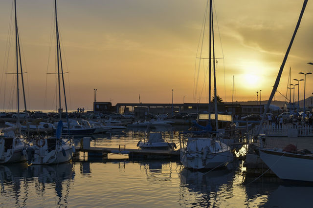 Tranquil scene of sailboats docked at a harbor in Sicily during a golden sunset, reflecting on the water.