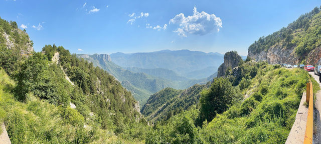Wide panorama of a green gorge in the Albanian mountains, with the mountain road and parked cars along the right-hand edge