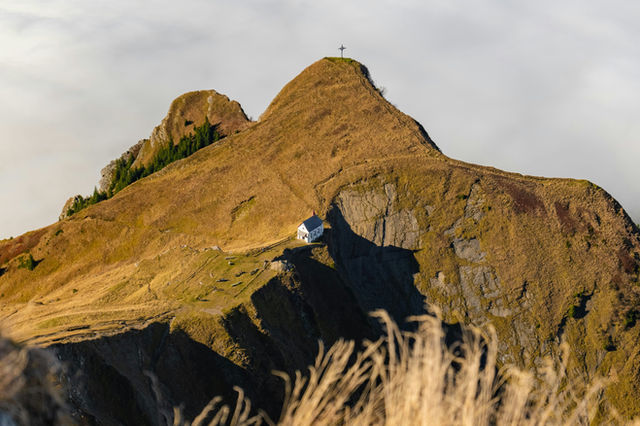 A small white building sits on a grassy mountain peak.