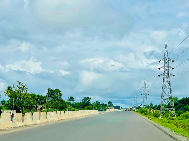 This image captures the highway leading to Roberts International Airport, flanked by lush greenery and tall power lines. The open road stretches into the distance under a cloud-filled sky, highlightin