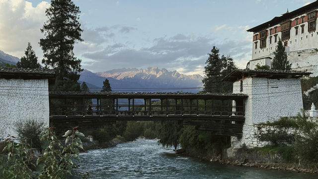 A traditional wooden bridge leads to Paro Dzong, spanning a gentle river beneath the soft glow of dusk, with distant Himalayan peaks completing the tranquil scene.