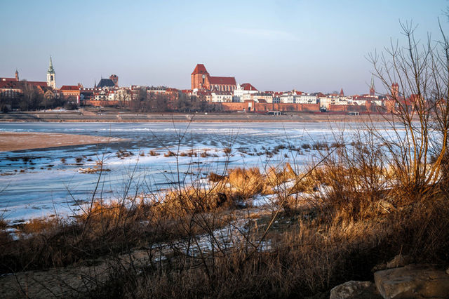 A scenic winter view of the historic Toruń cityscape across an ice-covered Vistula River.