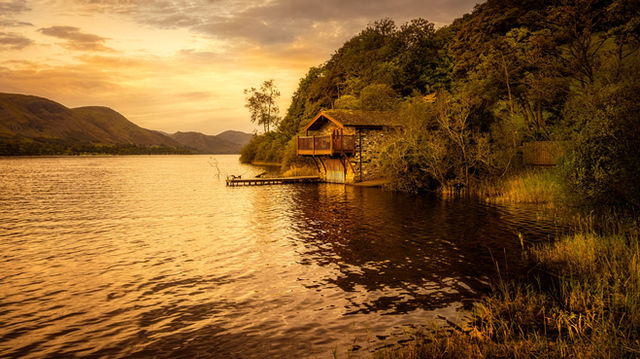 A wooden house by a lake reflecting a stunning orange sunset, surrounded by lush trees.