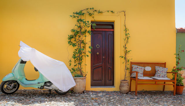 Colorful scene with a covered scooter and wooden bench against a yellow wall in Bosa, Sardinia.
