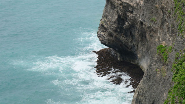 Lovers' Cliff巴厘岛西南海岸乌鲁瓦图神庙的壮丽景象。70 米高的悬崖绝壁直插波涛汹涌的印度洋，层理分明的岩壁与巨大礁石是千万年海浪侵蚀的杰作。湛蓝海水与灰白色岩壁形成强烈对比，白浪拍岸的瞬间充满了原始的力量感。这里不仅是守护巴厘岛的印度教圣地，更是以绝美日落闻名的旅行胜地，完美融合了自然的鬼斧神工与人文的精神信仰。
