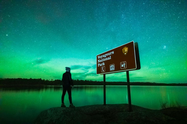 Photograph of person standing next to brown national park sign at night with northern lights.