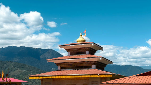 A beautifully crafted temple in Thimphu, Bhutan, rises against a backdrop of mountains and clouds, its golden stupa and layered roof showcasing the grace and depth of Bhutanese spiritual architecture.