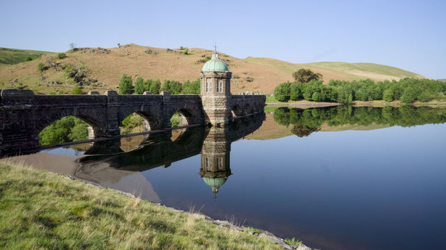 A scenic view of a historic stone bridge reflecting in a calm lake under a clear sky.