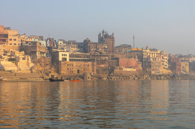 🛶 Ganges Riverfront at Varanasi – India This scenic image presents a tranquil view of the Ganges River with a hazy cityscape of Varanasi stretching along the horizon. The water appears calm and silver