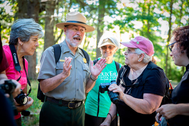 A park ranger in uniform  appears to lead a spirited discussion with a group of visitors in a wooded
