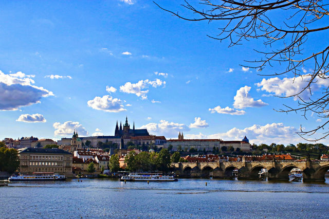 Bajo un cielo despejado salpicado de nubes, el Castillo de Praga se alza majestuosamente sobre la ciudad, con el emblemático Puente de Carlos cruzando el río Moldava en primer plano. Esta imagen captu