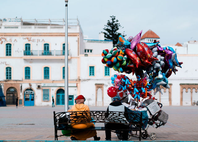 A vibrant balloon seller in Essaouira's town square, Morocco, with colorful buildings in the background.
