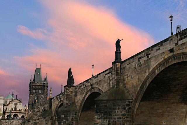 The photograph captures the historic Charles Bridge, one of Prague’s most iconic landmarks, bathed in the warm hues of sunset. The baroque statues rise majestically above the stone arches, while the s