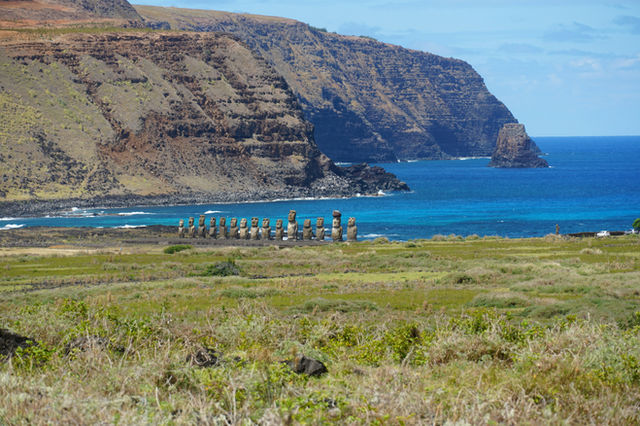 Moai statues stand on a grassy field by the ocean.