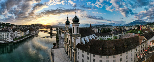 This stunning panorama captures Lucerne, Switzerland, in the breathtaking transition between blue and golden hour. The majestic Jesuit Church dominates the foreground with its iconic twin green domes,