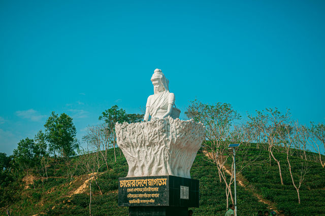A white statue of a female tea garden worker stands prominently against a vibrant, clear blue sky. The sculpture, depicting a woman with a harvesting basket, rests on a black pedestal with Bengali tex