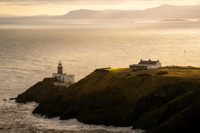A picturesque scene of a lighthouse and a house perched on a grassy cliff, with the sun setting over the vast ocean.