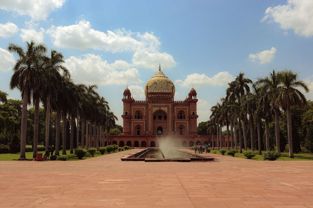 A scenic view of Safdarjung Tomb in New Delhi with palm trees and a fountain under a bright blue sky.