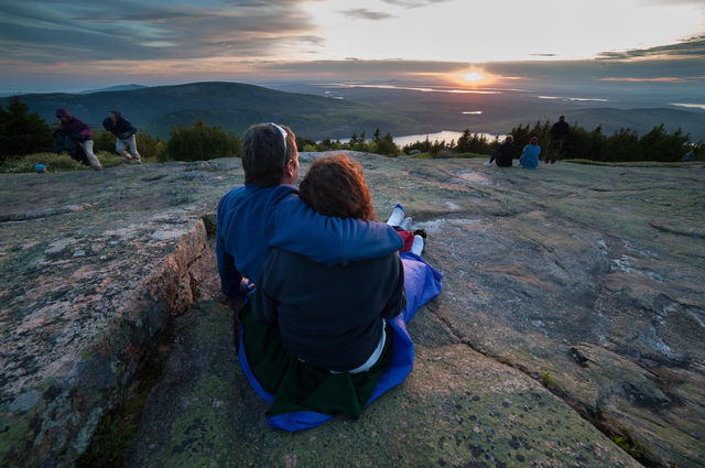 While seated on a blanket on a patch of mountaintop granite, a man and woman clasp their arms around