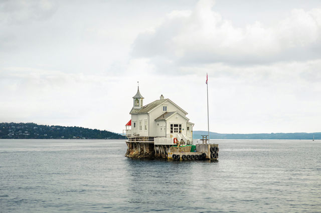 The historic Dyna Fyr lighthouse stands as a white wooden sentinel amidst the vast, gray expanse of the Oslofjord. Perched on a tiny rock outcrop, this charming example of traditional Norwegian mariti