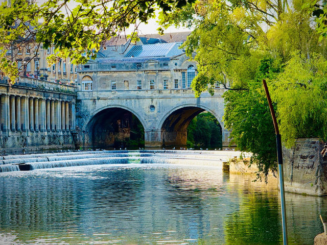 Scenic view of historic stone bridge in Bath, UK, reflecting in serene river surrounded by lush greenery.