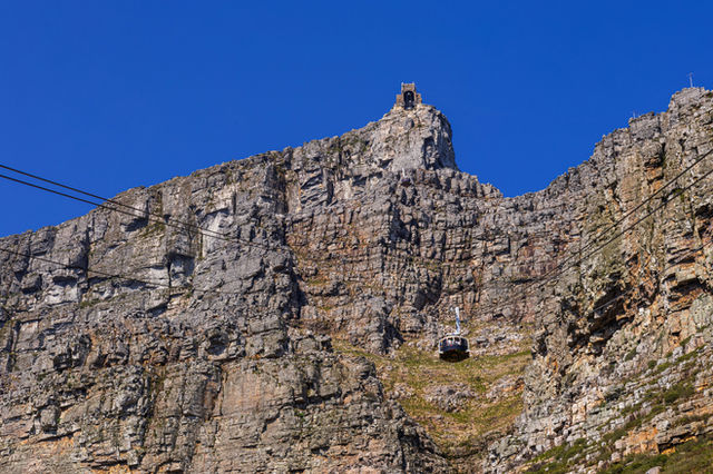 🚡 Cable Car Ascending Table Mountain, Cape Town, South Africa The image showcases a cable car gliding up the rugged face of Table Mountain, suspended in midair against the steep, rocky slope. Thick st