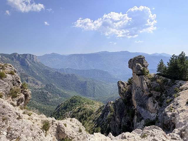 A limestone pinnacle stands above a forested gorge in the Albanian mountains, ridge after ridge fading into summer haze