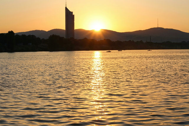 Esta fotografía muestra un cálido atardecer sobre el Río Danubio, con el sol ocultándose tras las colinas y reflejándose en el agua suavemente ondulada. La silueta de la moderna DC Tower 1 se eleva co