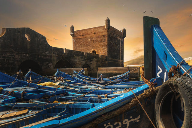 A scenic view of the historic Essaouira fortification with blue boats at sunset, capturing Moroccan culture.
