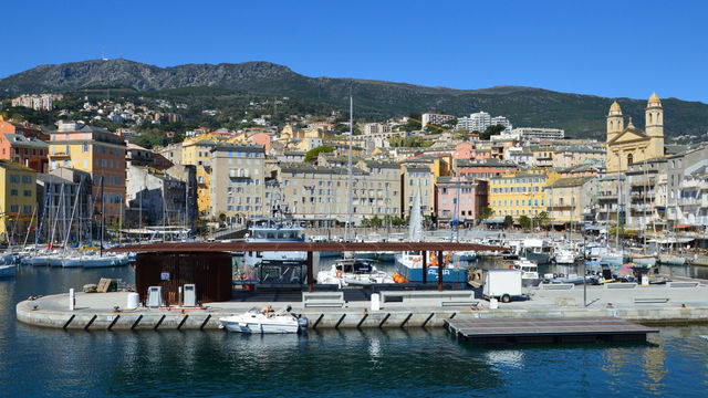 Picturesque harbor scene in Bastia, Corsica, featuring colorful buildings and mountain backdrop.