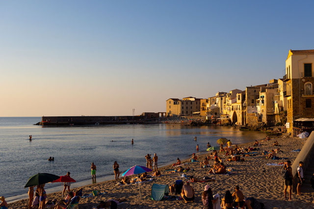 People enjoying a summer evening on the beach in Cefalù, Sicily, with historic buildings nearby.