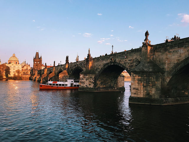 Scenic view of the Charles Bridge and iconic Prague landmarks during sunset with a boat on the Vltava River.