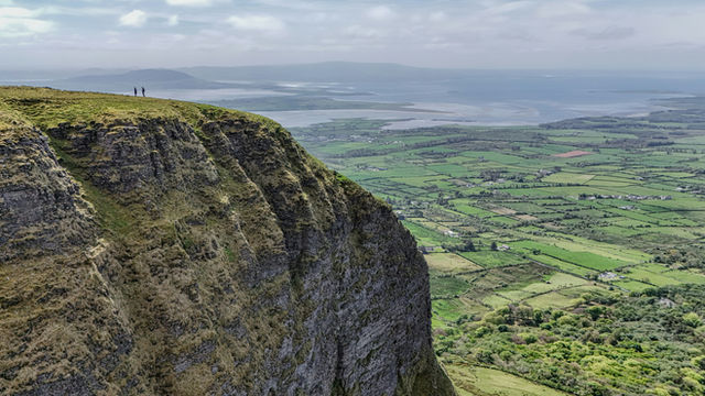 Two hikers stand on the edge of Benbulben, taking in the view. In the foreground, the mountain’s rugged texture contrasts with the vast landscape of green fields and water stretching out below.