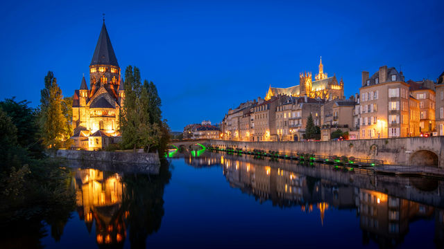 Metz by Night: A View from the Moselle. Located in northeastern France, Metz blends Roman, Gothic, and Germanic influences into a unique architectural mix. On the left, the illuminated Temple Neuf sit