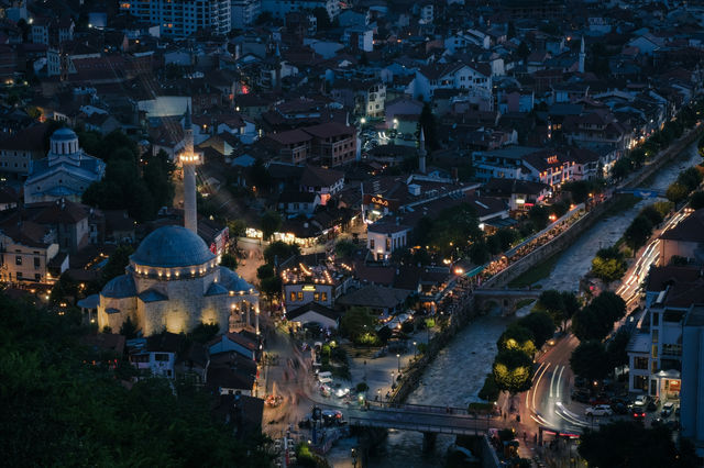 Prizren, a city where light and history intertwine. As dusk falls, the warm glow of streetlights and the illuminated mosque breathe life into the historic heart of the city. The winding river, stone b