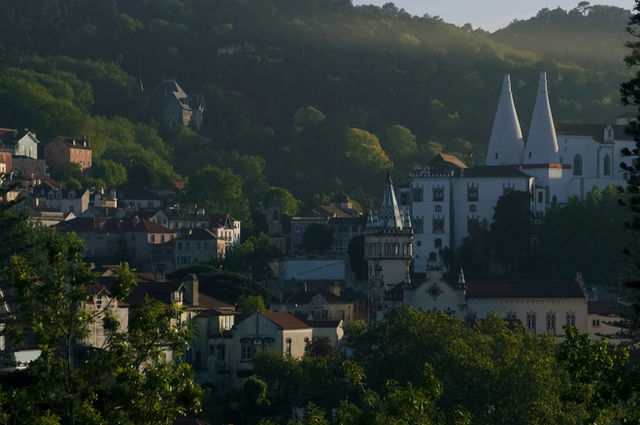 Sintra;-village,an;-Autumn;-morning;-fog;-mist;mystery;-mystical; A picturesque hillside town surrounded by lush green hills, featuring a prominent white historic building with two conical towers risi