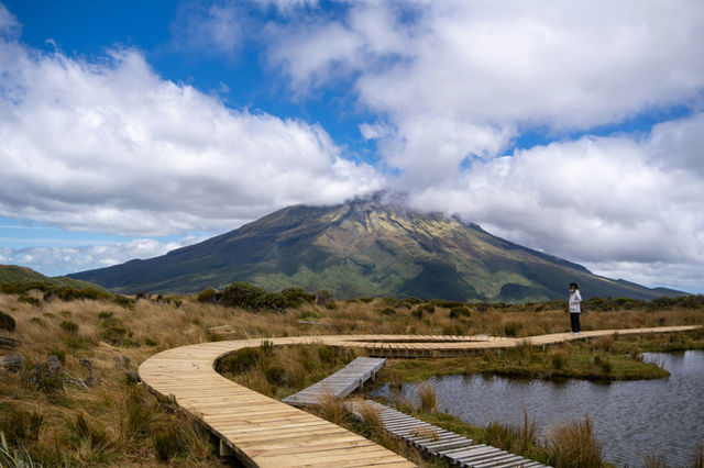 A lone person walks on a boardwalk near a mountain.