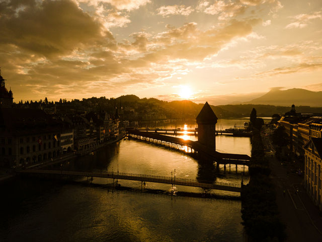 Experience the ethereal beauty of Lucerne, Switzerland, as the first light of dawn spills across the iconic Chapel Bridge and its historic Water Tower. This breathtaking golden hour scene captures the
