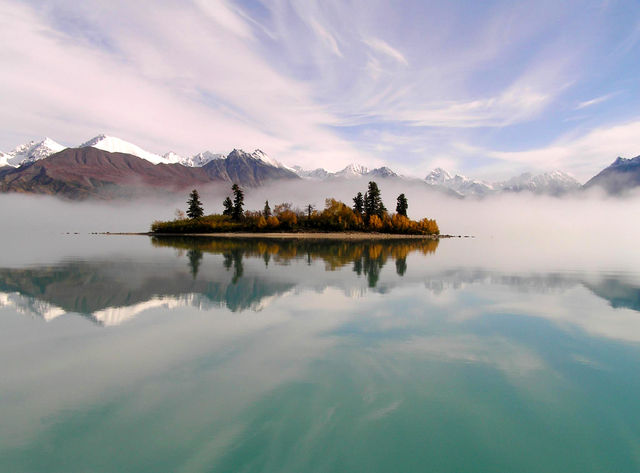 A colorful sky, an island with spruce trees, and mountains in the background reflect in a calm lake