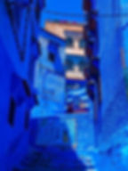 A narrow alleyway in Chefchaouen, Morocco, showcases vibrant blue walls under the bright daylight, with intricately designed window grilles and a colorful tapestry adding to the charm of the city known for its iconic blue-hued buildings.