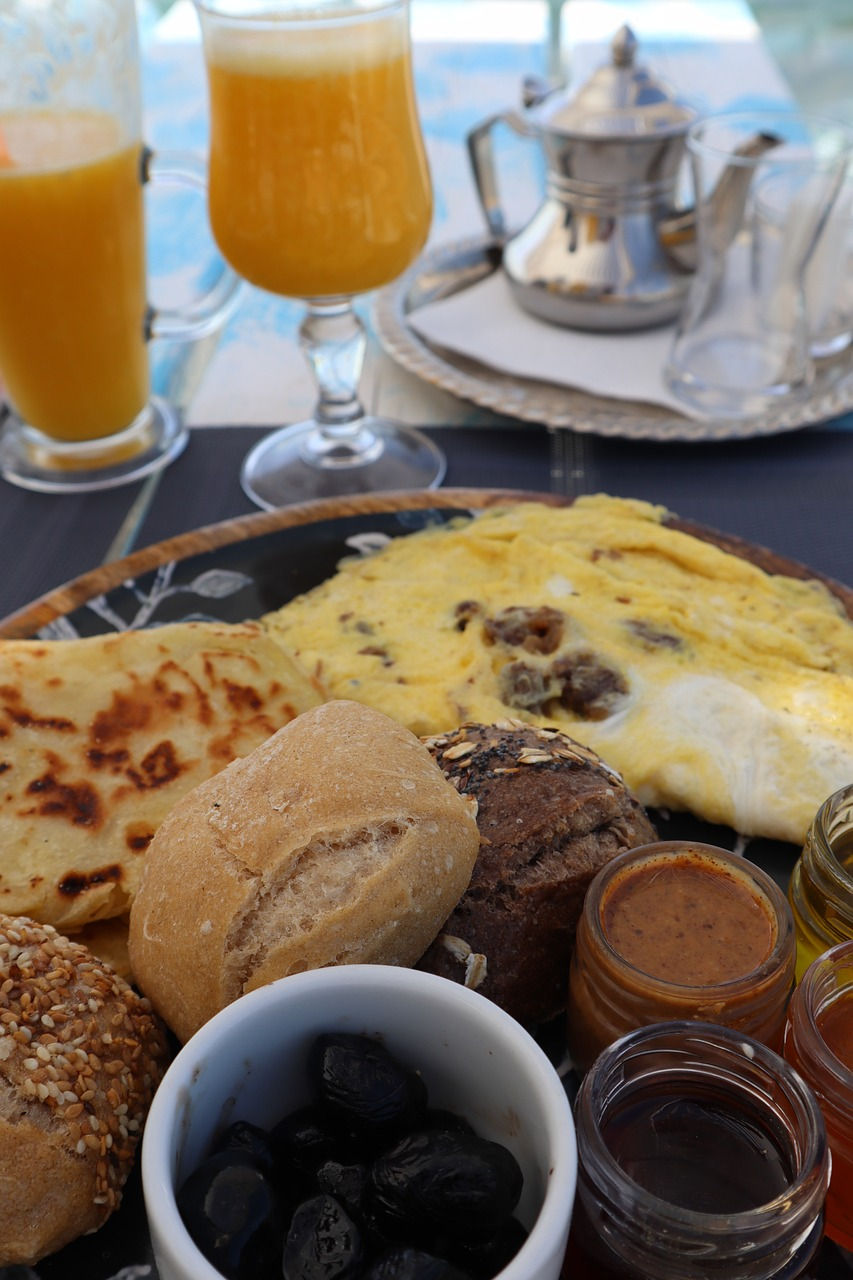 A sumptuous Moroccan breakfast spread featuring assorted breads, black olives, a flavorful khlea omelette, and a selection of traditional jams and honey, accompanied by a teapot ready to pour refreshing mint tea.