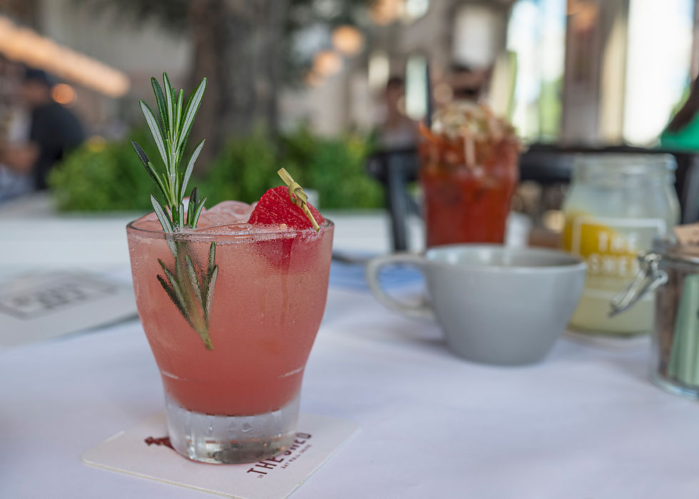 Pink cocktail with rosemary and strawberry garnish on a table. Blurred background with a drink, cup, and jar. Bright and inviting setting.